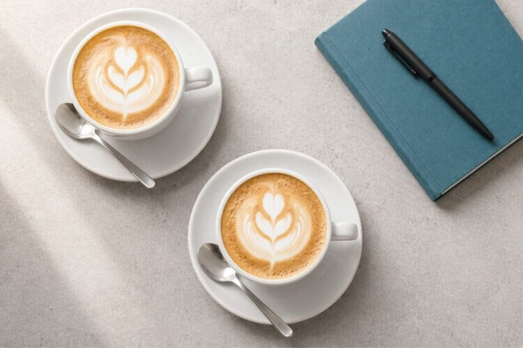 Twee cappuccino's met latte art naast een teal notitieboek en pen als symbool voor een vakinhoudelijk gesprek, een koffiedate, loopbaanoriëntatie en gesprekstraining. Loopbaanbegeleiding, boosttraject, sollicitatie